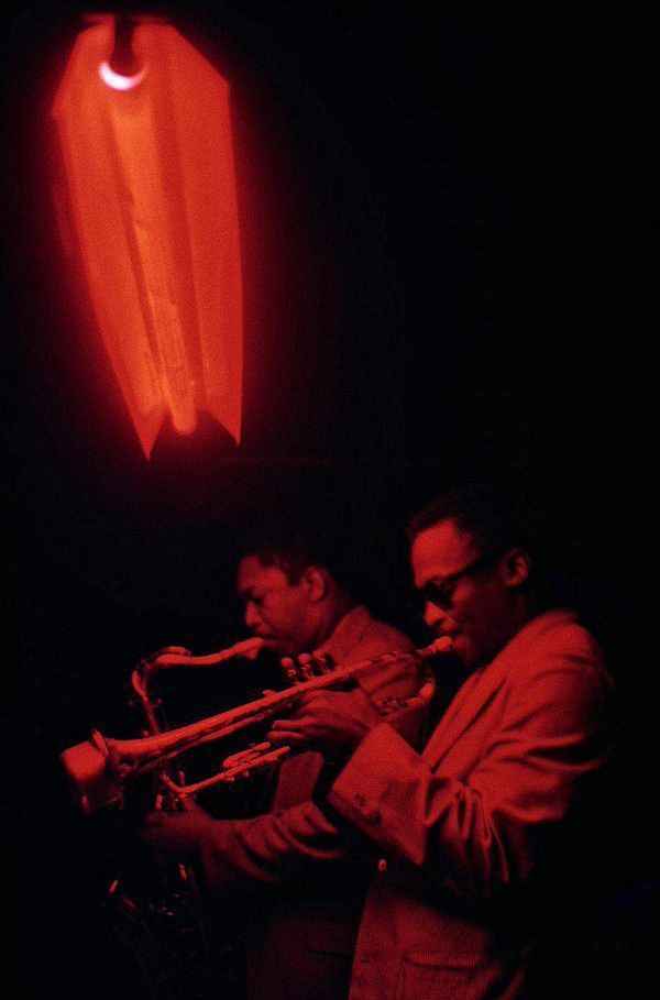 two men playing instruments in front of a bright orange light on a black background with an object hanging from it's side