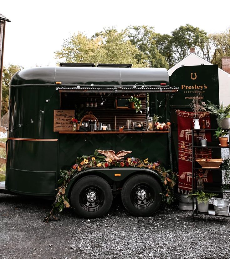 a green food truck parked in front of a wooden building with flowers on it's side