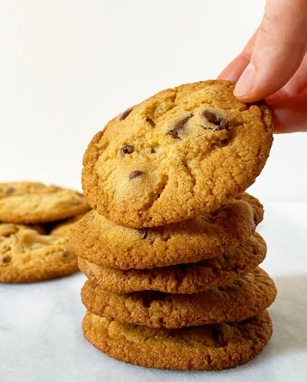 a hand picking up a chocolate chip cookie from a stack of cookies on a white surface