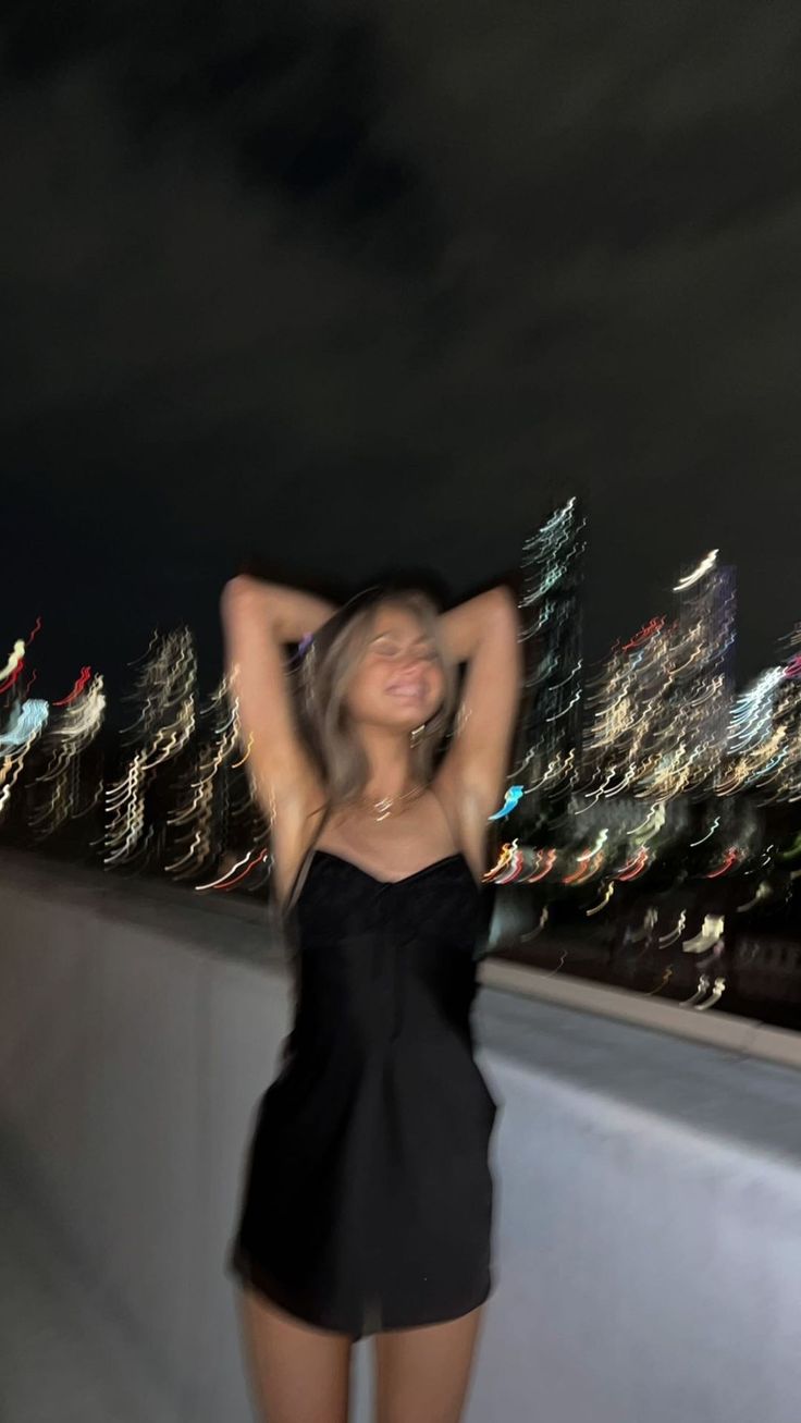 a woman standing on top of a roof next to a tall building at night with her hands behind her head