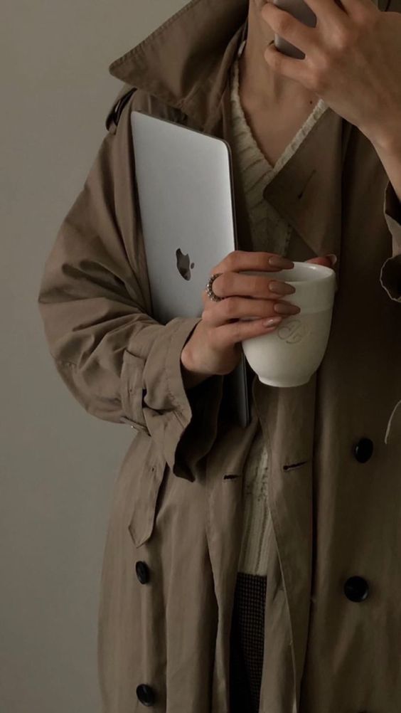 a woman in trench coat holding a coffee cup and an apple laptop computer with her right hand