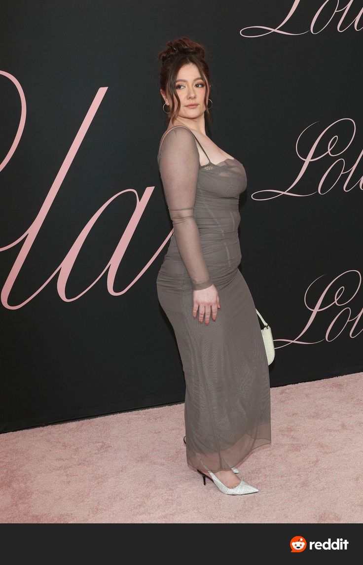 a woman in a grey dress standing on a pink carpet with her hand on her hip