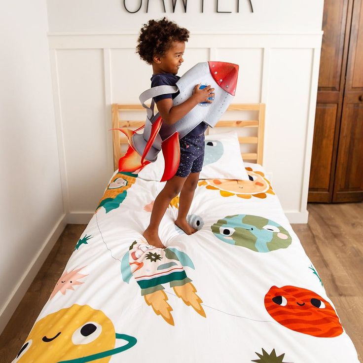 a little boy standing on top of a bed holding a toy rocket and looking at the camera