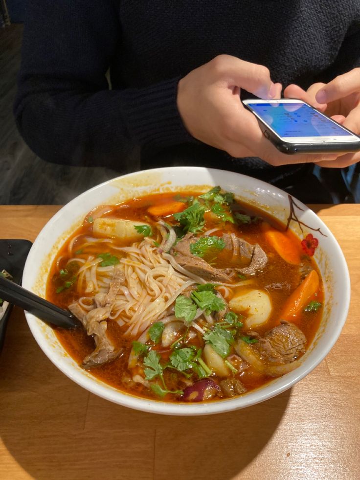 A white bowl of reddish broth sits on a tan-coloured wooden table. There are vibrant orange carrots, floating pieces of beef, and lots of noodles sitting in the glistening broth. The soup is garnished with greens. A black spoon leans on the inside of the bowl. Behind the soup is a man’s (my boyfriend’s) arm in a very dark sweater. He is holding his iPhone with both hands. Spicy Beef Pho, Winter Relationship, Pho Food, Boyfriend Date, City Downtown, Vietnamese Restaurant, Hits Different, Spicy Beef, Food Babe