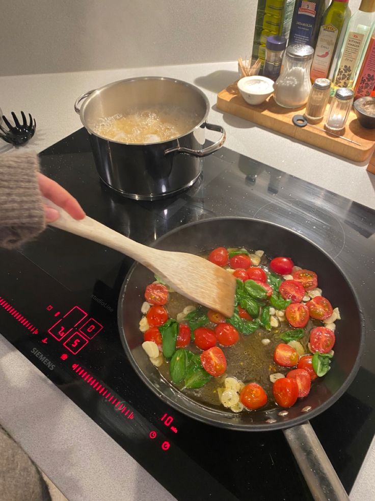 a person stirs tomatoes in a skillet on the stove with a wooden spoon