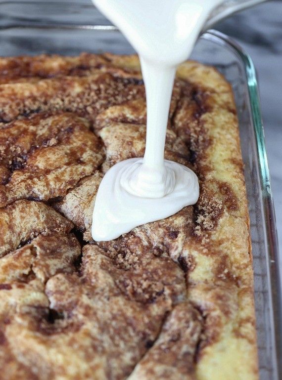 a white liquid pours into a square casserole dish filled with cinnamon rolls