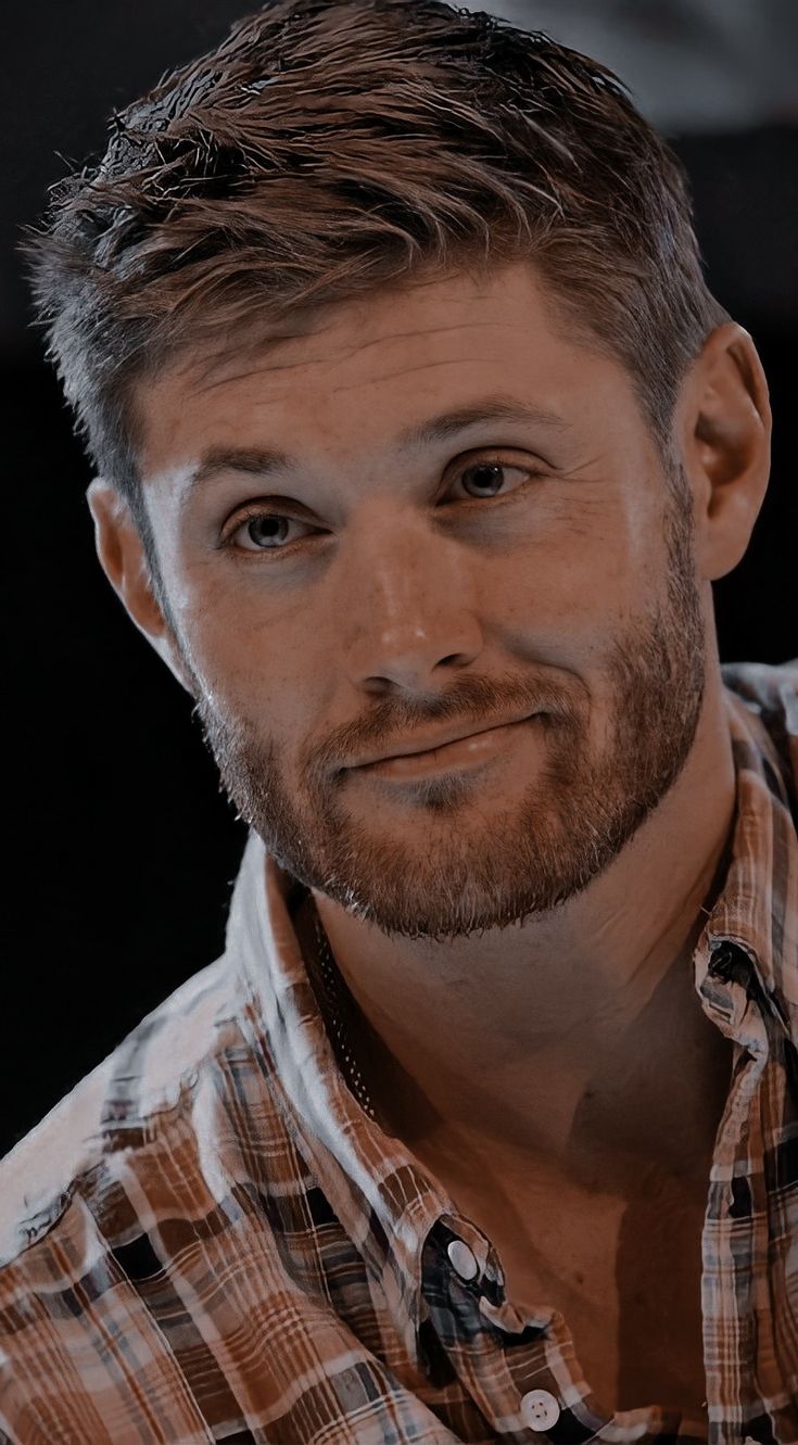 a man with a goatee and plaid shirt looks at the camera while he is smiling