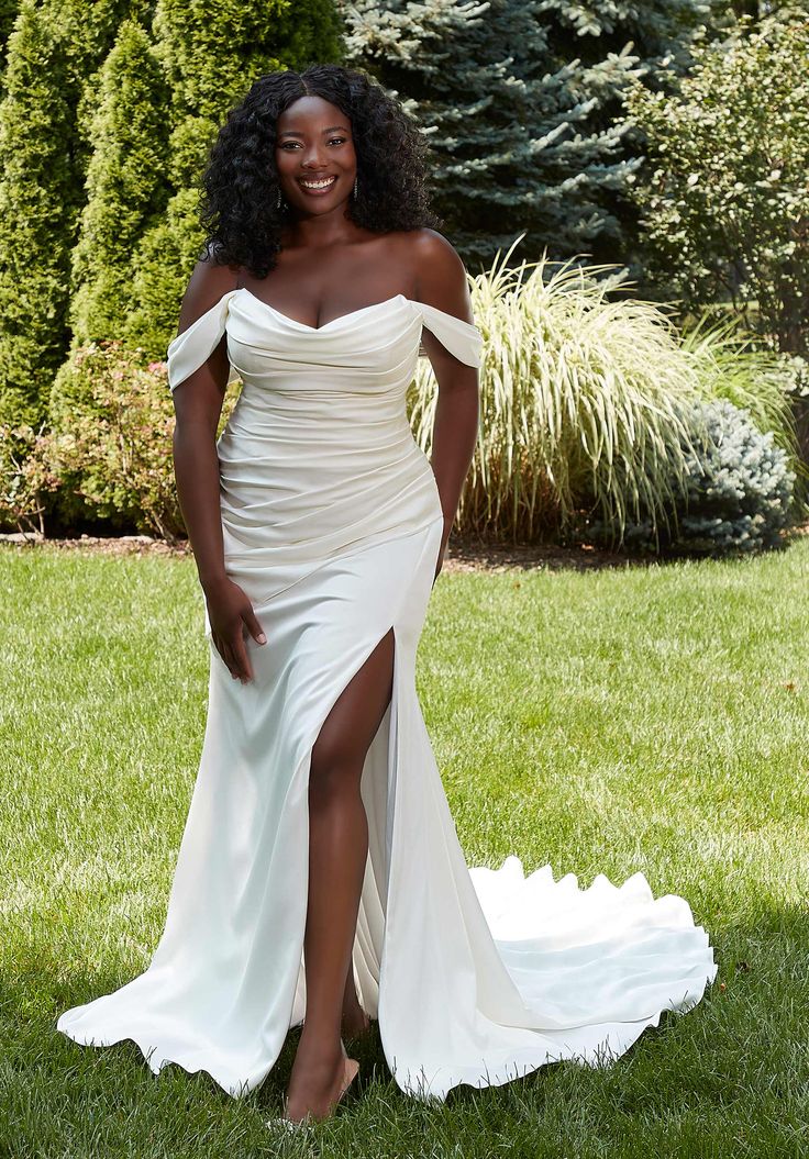 a woman in a white dress is posing for the camera on some grass with trees and bushes behind her