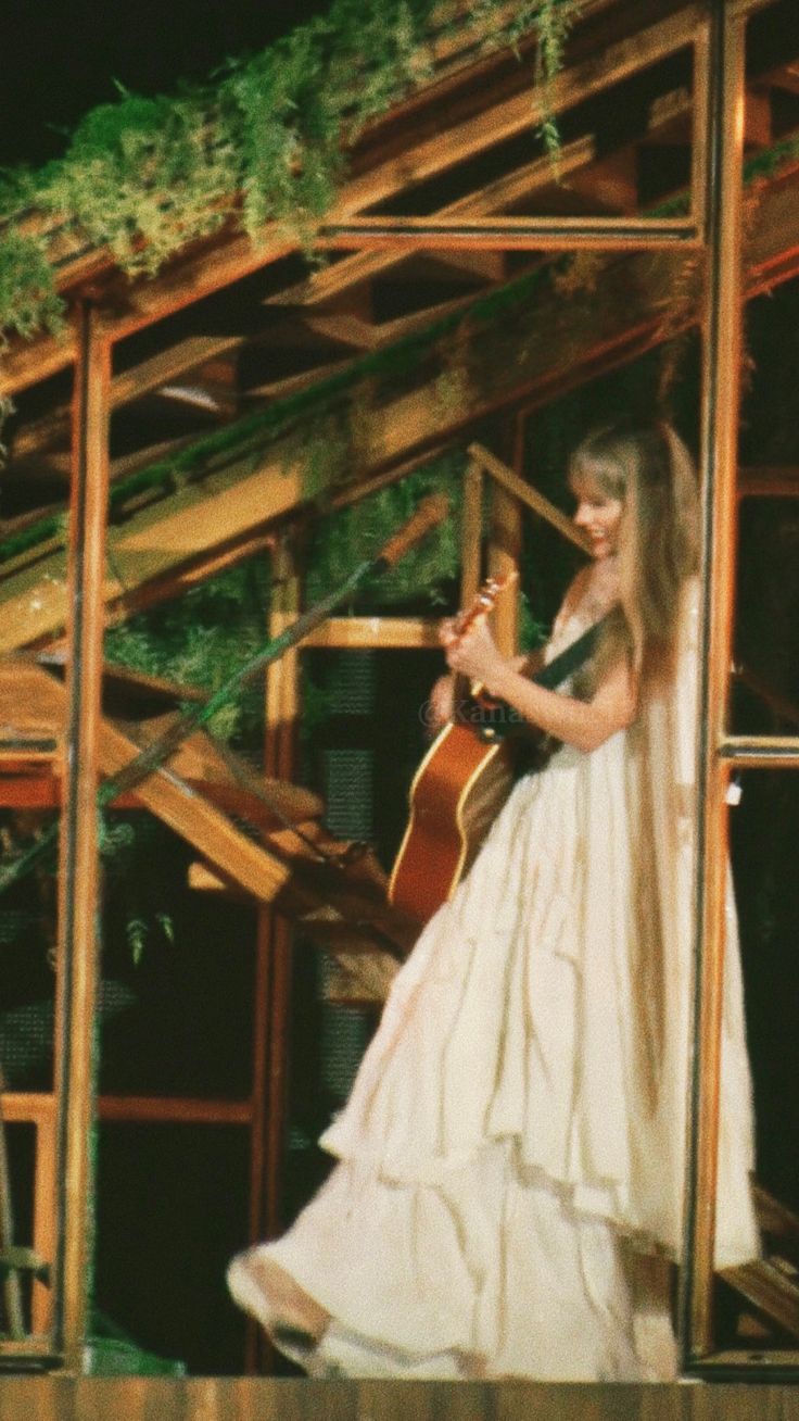 a woman in a white dress holding a guitar and standing next to a wooden structure