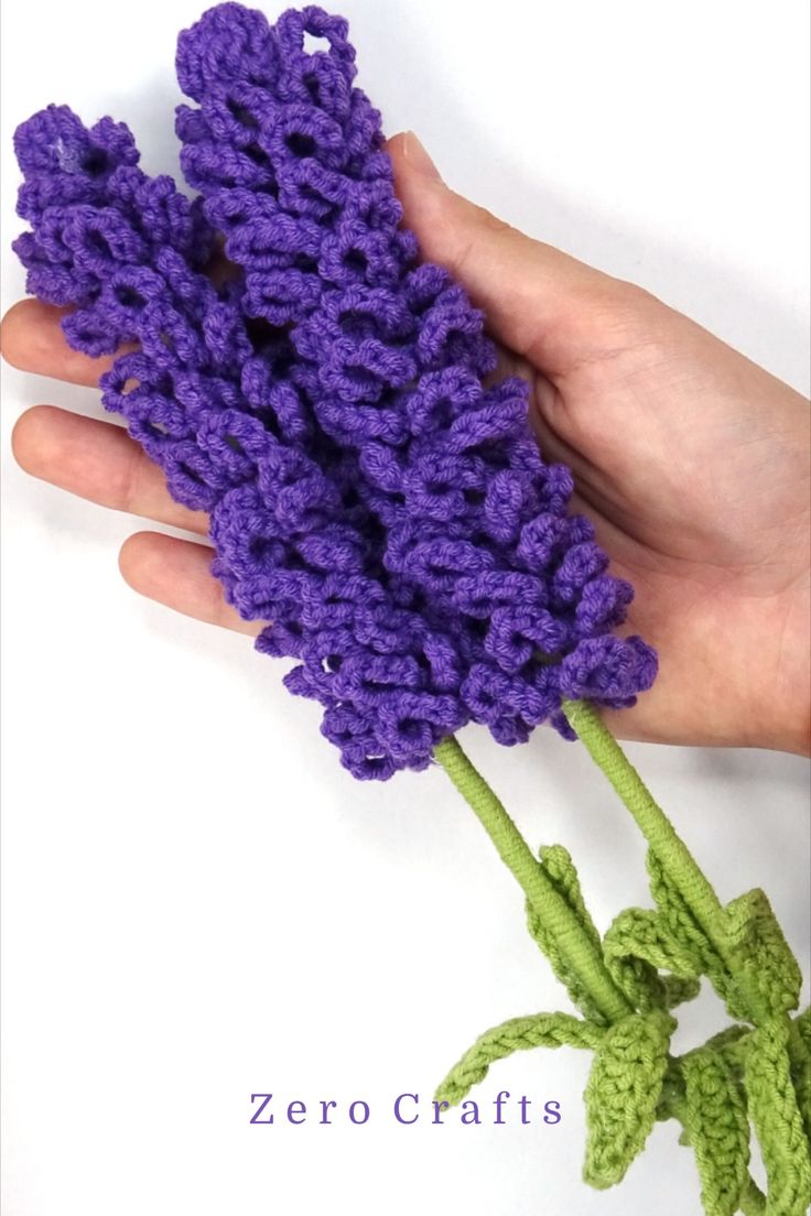 a hand holding a purple crocheted flower on top of a white surface with the words zero crafts written below it