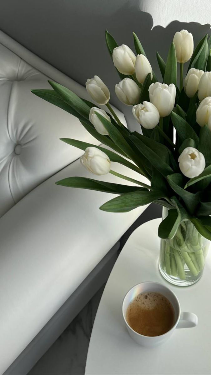 a vase filled with white tulips sitting on top of a table next to a cup of coffee