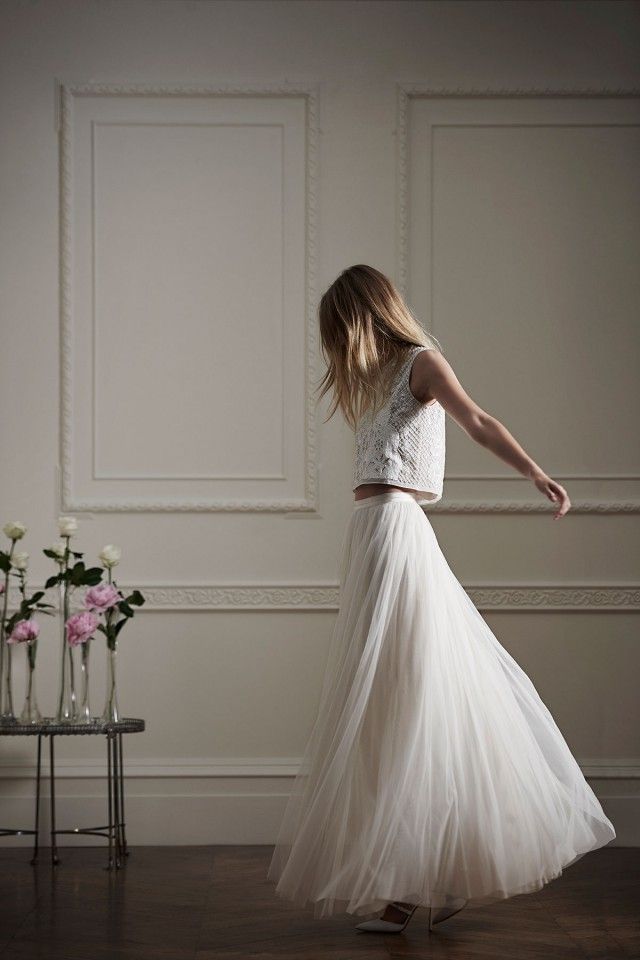 a woman in a long white skirt and crop top is standing by a table with flowers