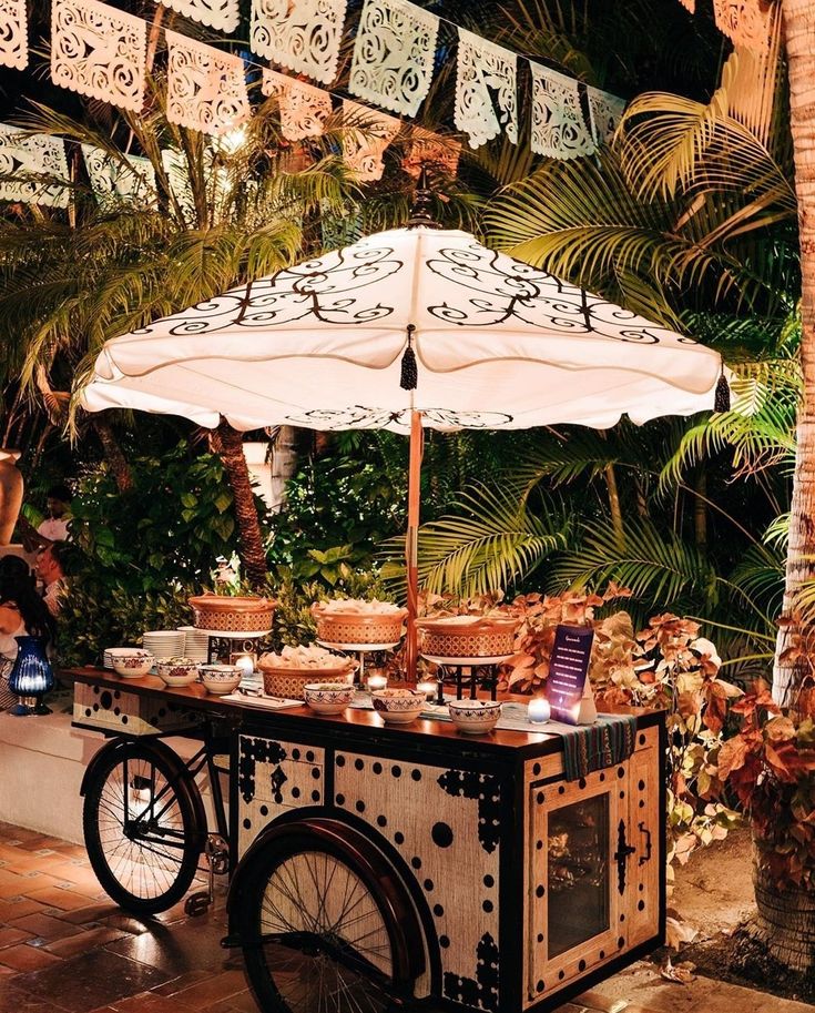 an outdoor food cart is set up with umbrellas and potted plants in the background