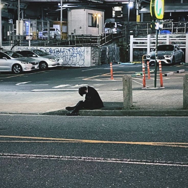 a person sitting on the side of a road next to a parking lot at night