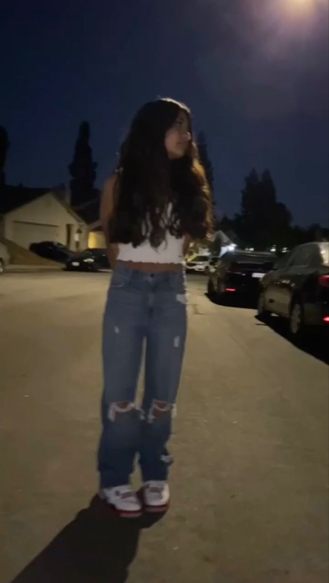 a woman standing in the middle of a parking lot at night with her hand on her hip