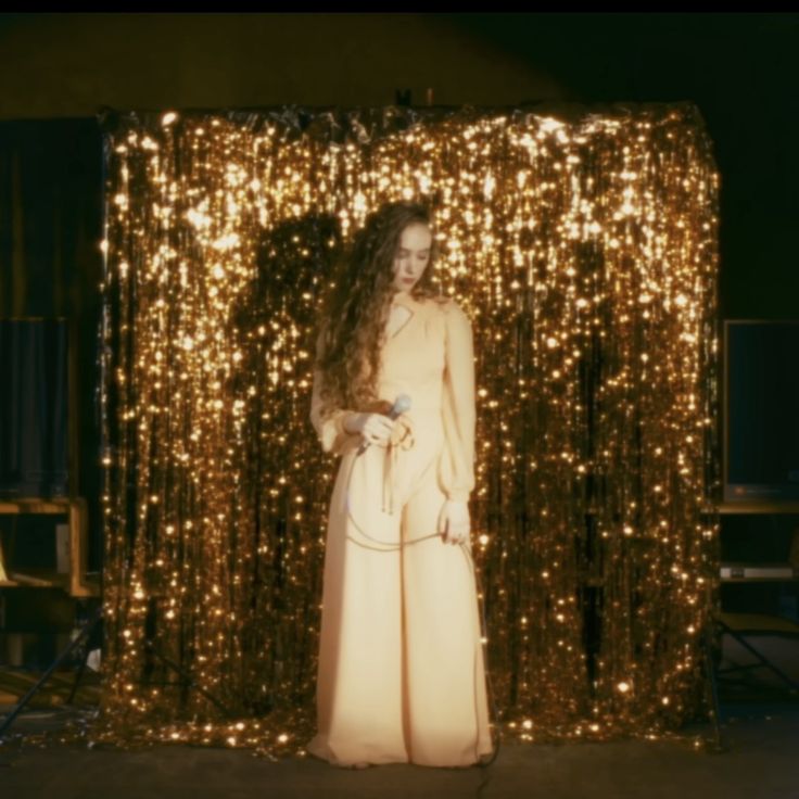 a woman standing in front of a gold curtain with lights on it's sides