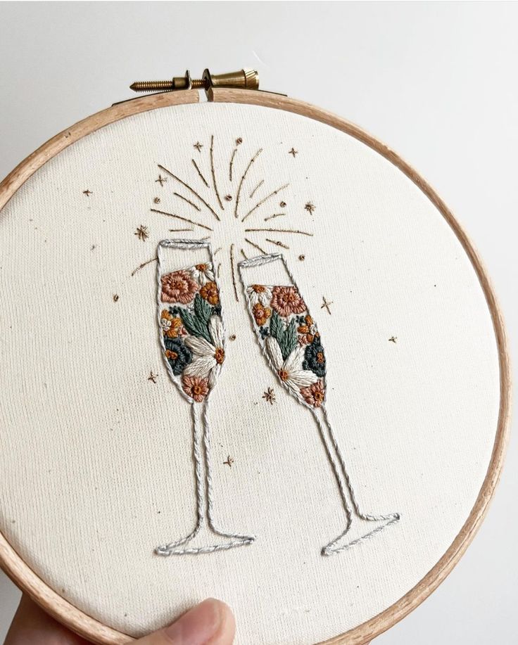 someone is holding up a cross stitch embroidery hoop with two champagne glasses on it and fireworks in the background