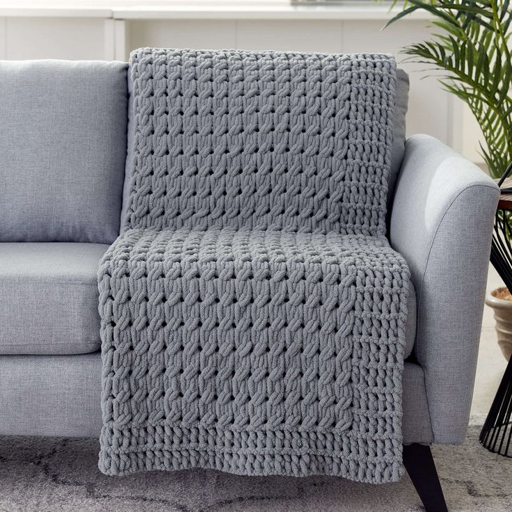 a gray couch with a crocheted blanket on it's back in front of a potted plant