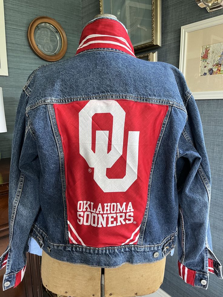 Boomer Sooner!  A vintage denim jean jacket is adorned with University of Oklahoma additions to create a one of a kind OU spirit jacket.  The collar, cuffs, and back are adorned with football logo material.  A unique and adorable one of a kind piece.  Size medium is available for shipping tomorrow .  Pre- order available for other team jackets in any size with up to 4 weeks creation time. SilkWarrior specializes in vintage jackets upcycled with designer scarves and fabric, creating unique and stylish pieces that stand out from the crowd. Each jacket is meticulously crafted to blend classic designs with modern trends, offering a one-of-a-kind fashion statement for the bold and adventurous. Located in Carthage, Missouri, SilkWarrior is the go-to destination for those who appreciate the art o Painted Jean Jacket Lsu, Football Jean Jacket Diy, Football Denim Jacket, Spring Denim Outerwear For College, Collegiate Cotton Outerwear For Spring, Long-sleeved Cotton Denim Jacket For College, Long Sleeve Cotton Denim Jacket For College, Casual Cotton Denim Jacket For College, Cotton Long Sleeve Denim Jacket For College