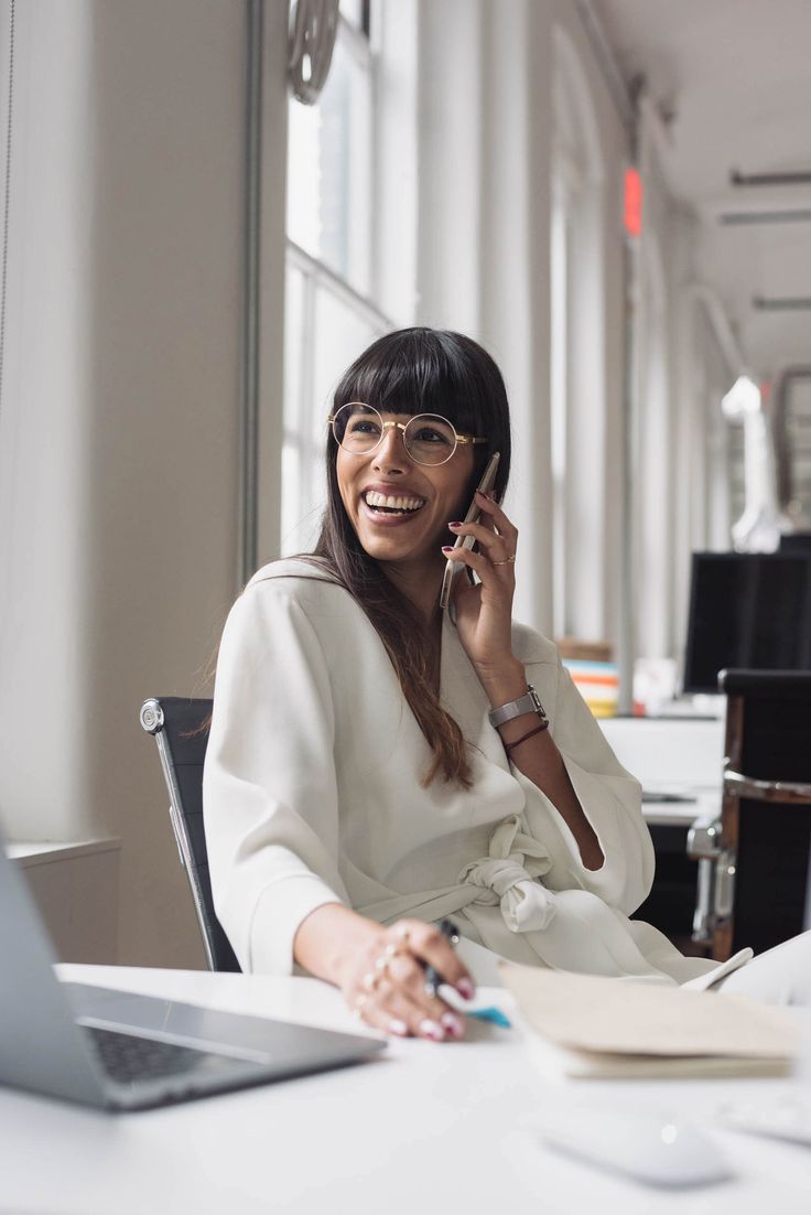 a woman sitting in front of a laptop computer talking on a cell phone and smiling