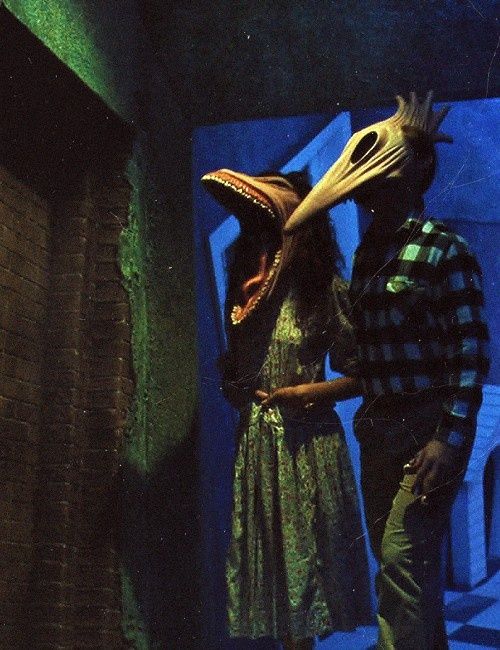 two people standing next to each other near a brick wall with a bird mask on their head