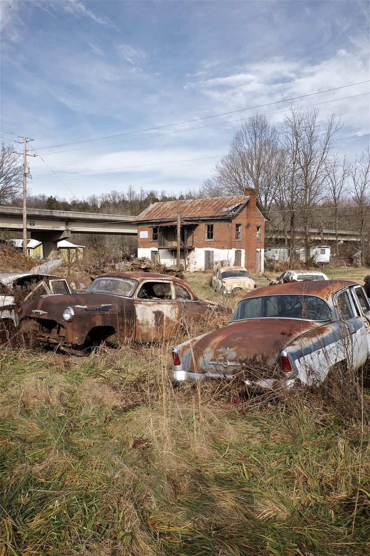 an old rusted out car sits in the middle of a field with other abandoned cars