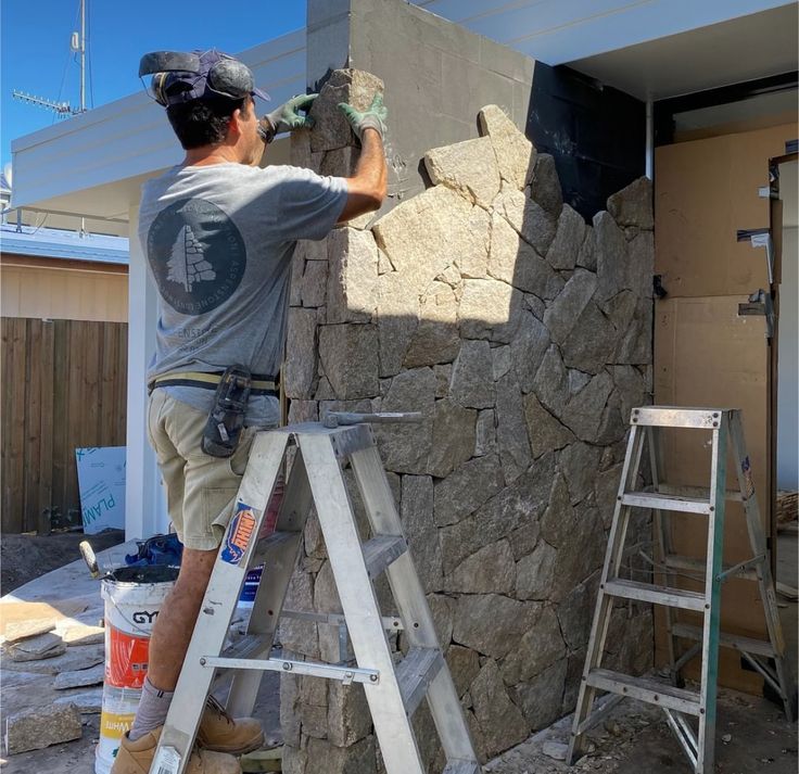 a man that is standing on a ladder near a rock wall and painting the walls