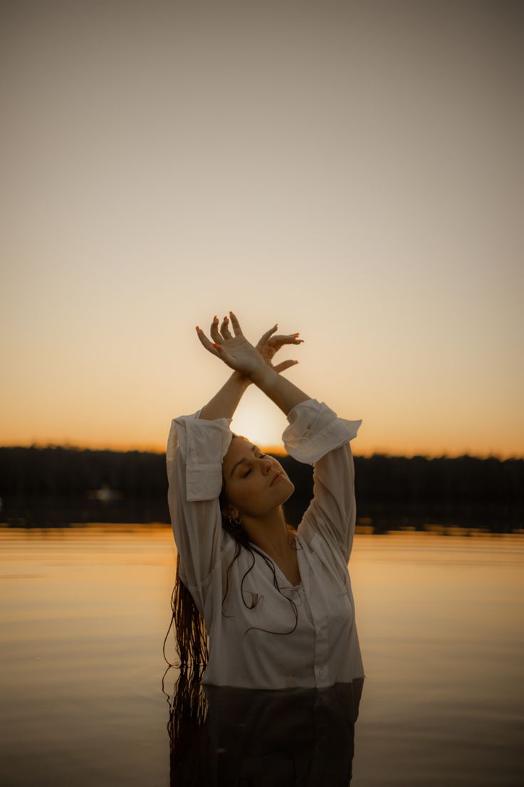 a woman standing in the water with her hands up to her head as the sun sets