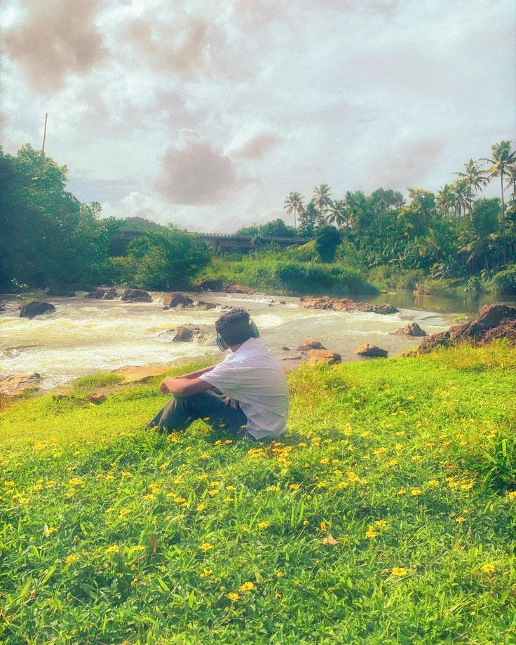 a man sitting on top of a lush green field next to a river under a cloudy sky