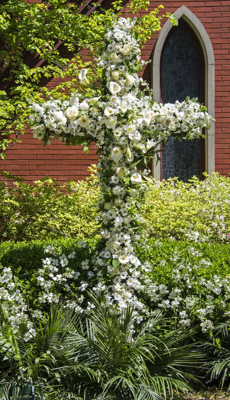 a white cross sitting in the middle of a garden next to a brick building with a stained glass window