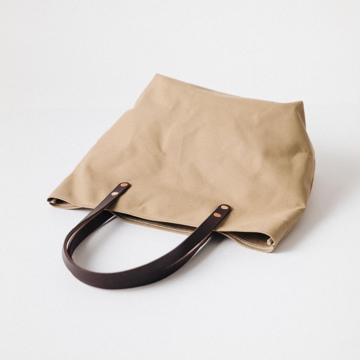 The Field Tan Canvas tote is a canvas and leather tote handmade in the KMM & Co. studio in Atlanta. Each one is meticulously crafted from heavyweight canvas and gorgeous full-grain leather. Field Tan Canvas is a type of cotton canvas known as duck cloth. It's one of the most durable types of cloth, and it's structured enough to stand up on its own. The Field Tan Canvas tote comes standard with Dark Brown handles. Canvas Shoulder Tote Bag With Rolled Handles, Canvas Bag With Leather Handles And Top Handle, Khaki Canvas Bag, Canvas Tote Shoulder Bag With Rolled Handles, Canvas Tote Bag With Rolled Handles, Cotton Canvas Top Handle Bag For Everyday Use, Everyday Waxed Canvas Shoulder Bag With Top Carry Handle, Everyday Use Cotton Canvas Top Handle Bag, Everyday Waxed Canvas Shoulder Bag With Top Handle