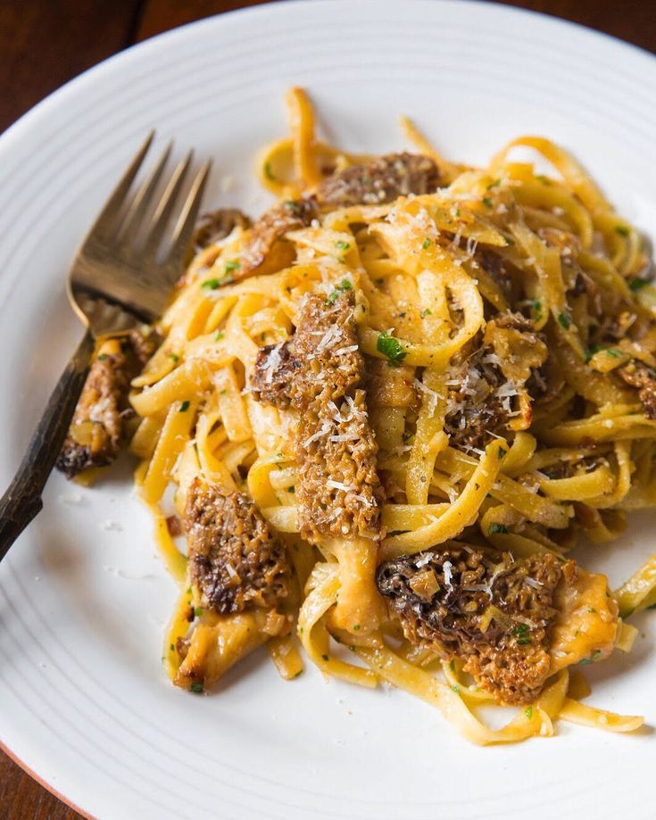 a white plate topped with pasta and meat covered in sauce next to a fork on top of a wooden table