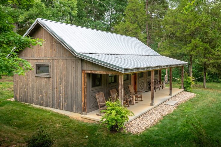 a small cabin in the middle of a wooded area with tables and chairs on the porch