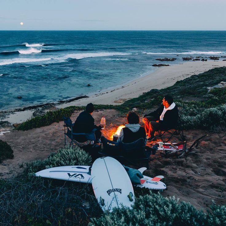 four people sitting around a campfire on the beach with surfboards in front of them