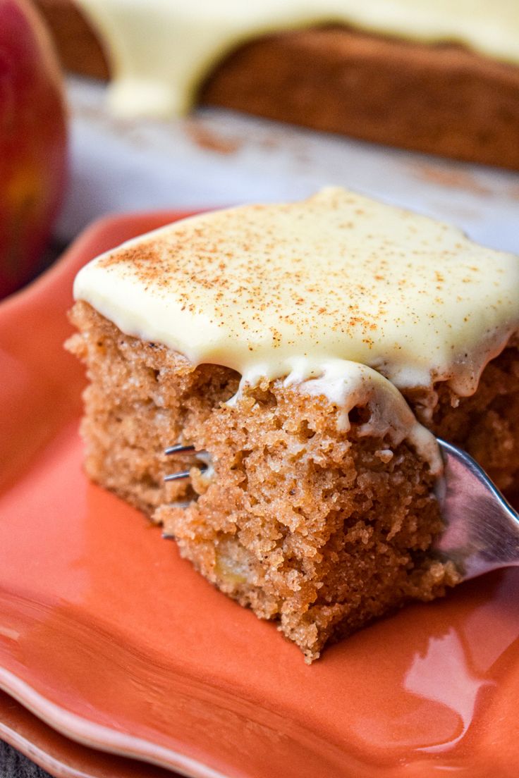 a piece of carrot cake with cream cheese frosting on an orange plate next to an apple