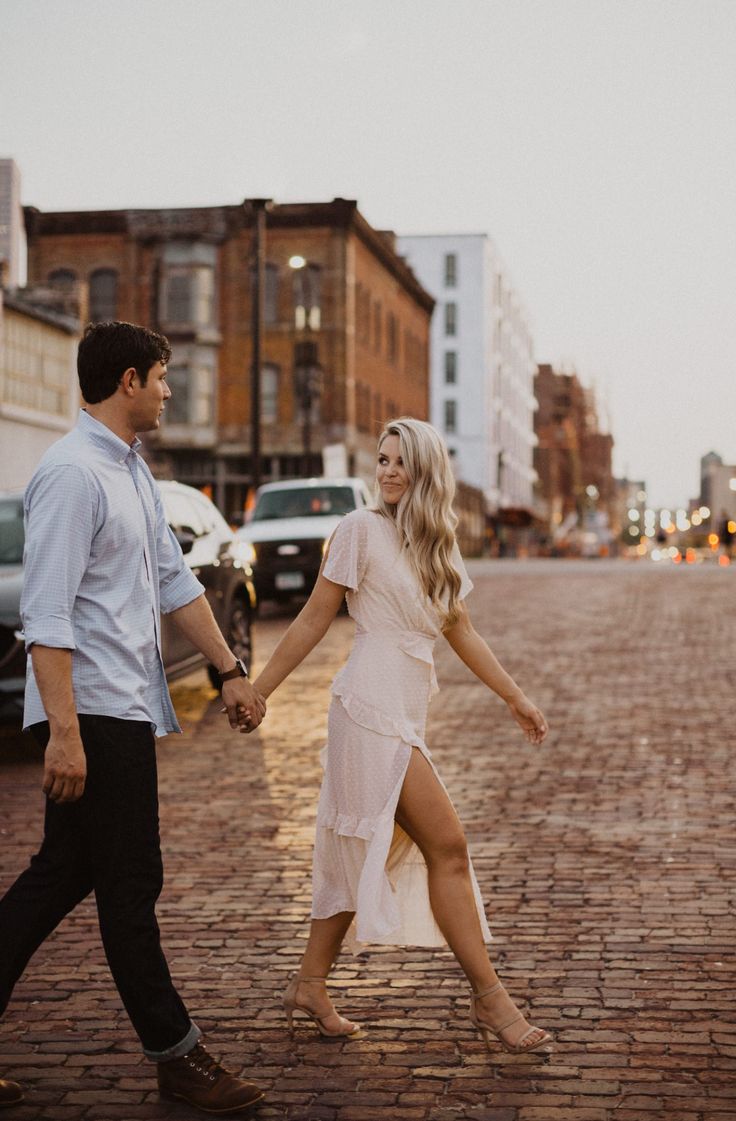 a man and woman holding hands walking down the street in front of parked cars on a cobblestone road