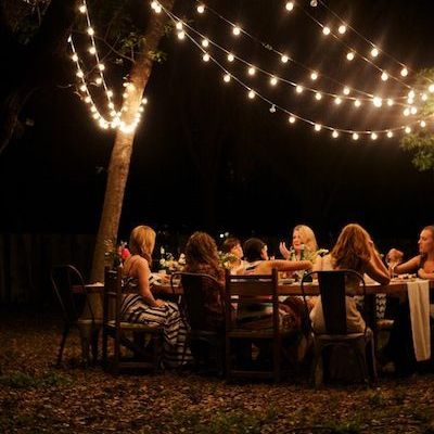 a group of people sitting around a dinner table at night with lights strung over them