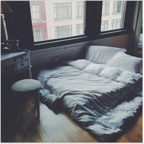 an unmade bed sitting in the middle of a room next to two large windows