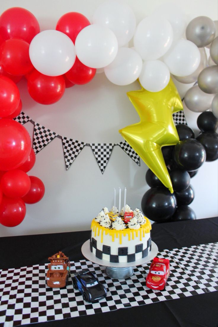 a birthday party with balloons, cake and cars on a checkerboard tablecloth