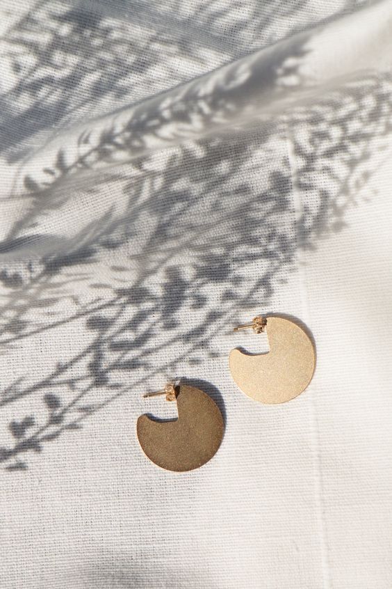 two pairs of gold earrings sitting on top of a white cloth covered table next to a plant