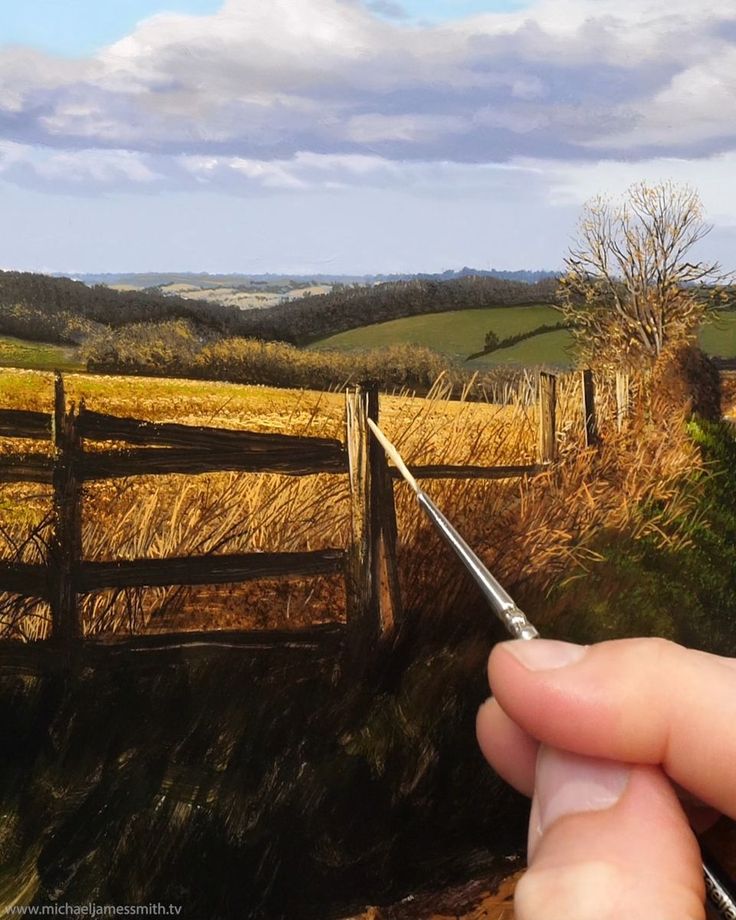 someone is holding a paintbrush in front of a painting of a fence and field