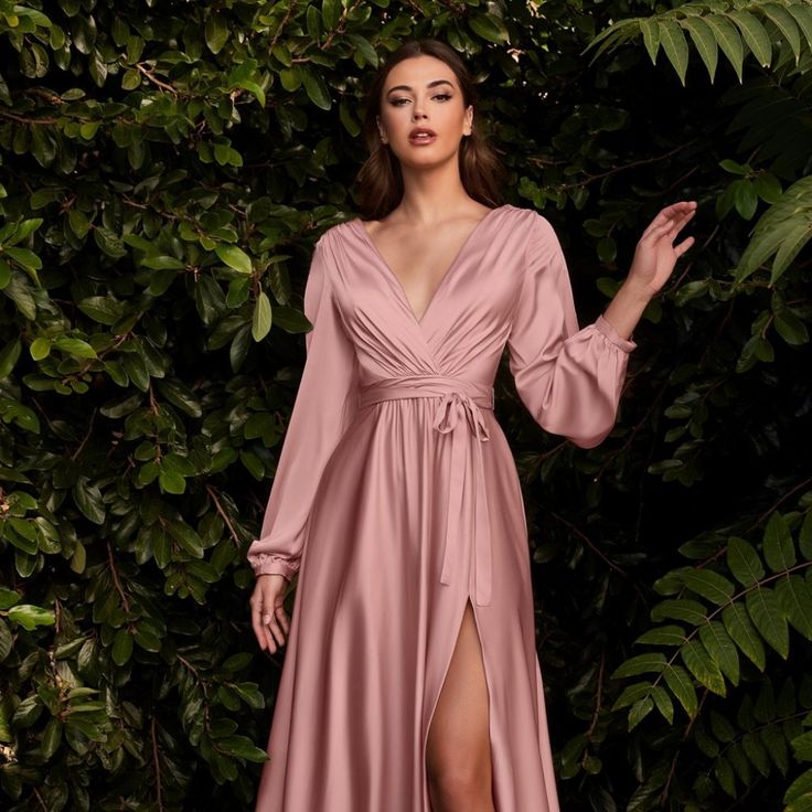 a woman standing in front of some trees wearing a pink dress with long sleeves and a thigh high slit