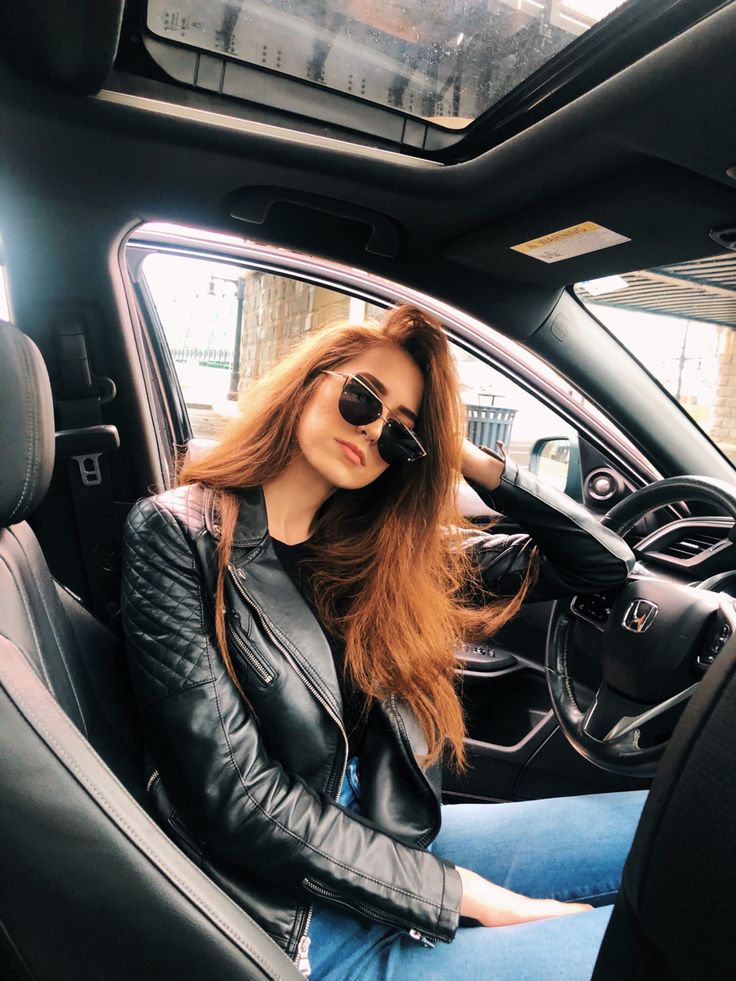 a woman sitting in the driver's seat of a car wearing sunglasses and a leather jacket