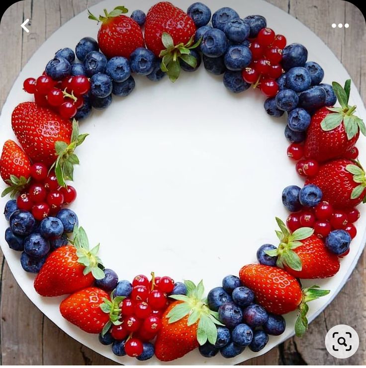 a cake decorated with strawberries and blueberries on a white plate next to a wooden table