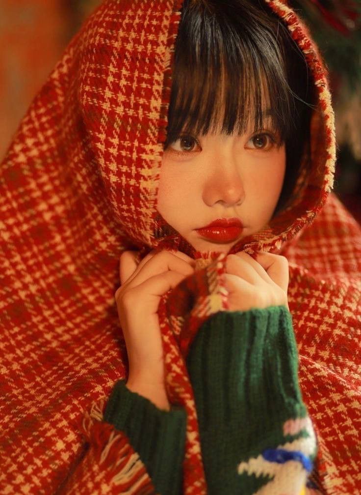 a young woman with dark hair wearing a red and green coat, holding her hands under her face