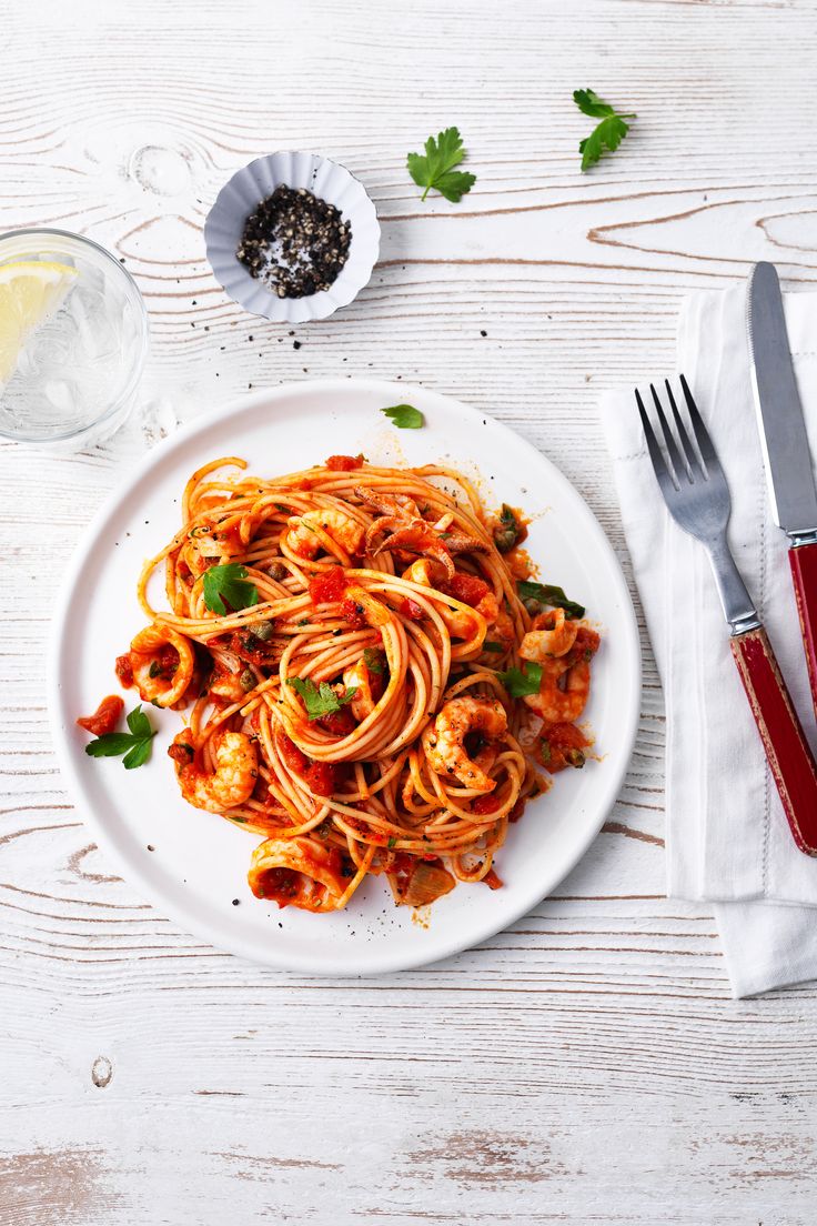 a white plate topped with pasta next to a fork and glass of water on top of a wooden table