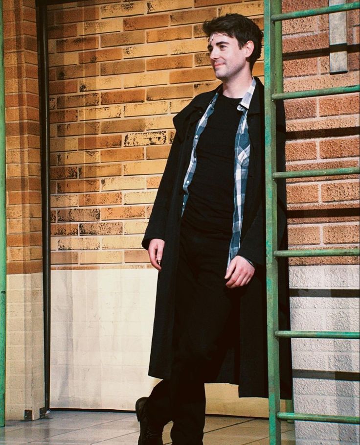 a man standing in front of a brick wall with a coat over his shoulders and boots on