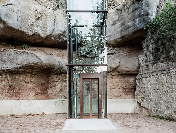 an entrance to a glass walled building in front of a rock wall with trees growing out of it