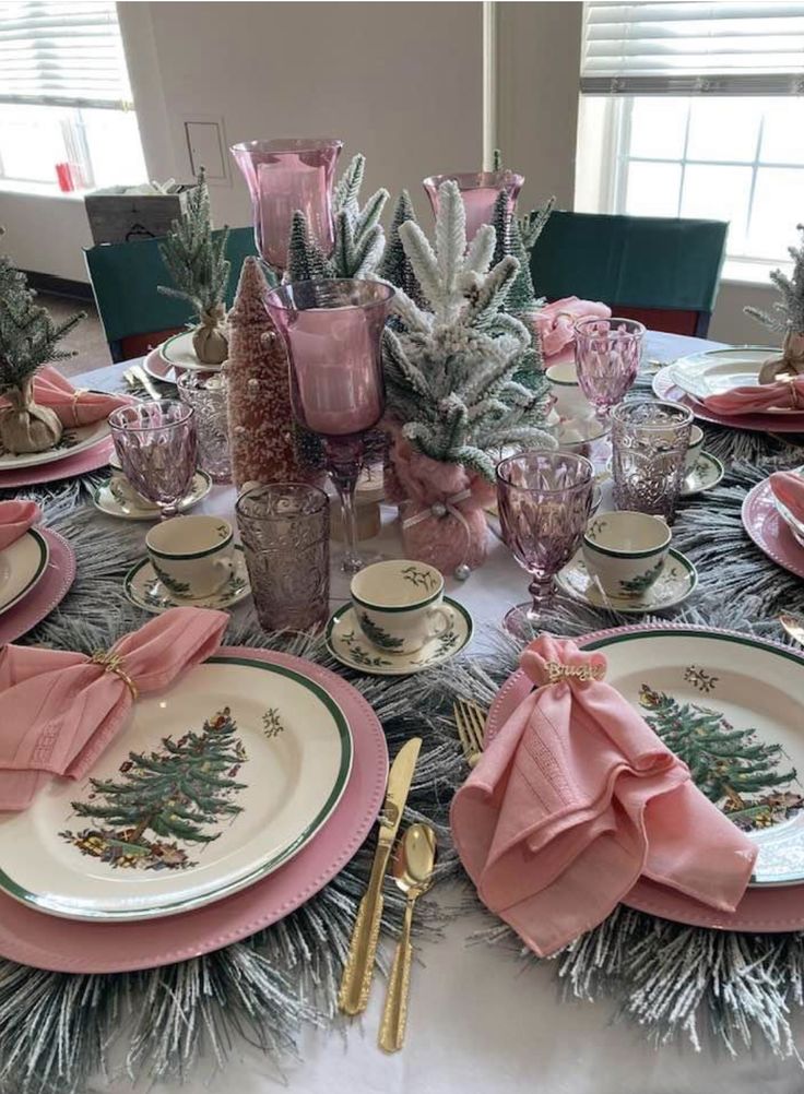 the table is set with pink and white dishes, silver place settings, pine trees, and other holiday decorations