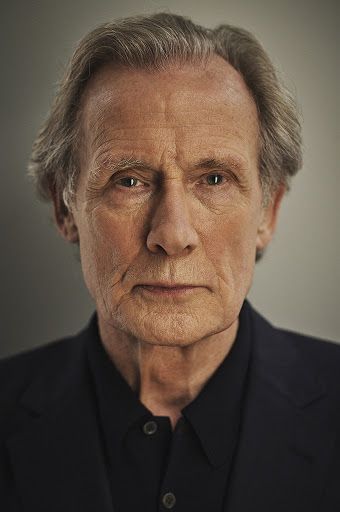 an older man with gray hair and black shirt looking at the camera while standing in front of a grey background